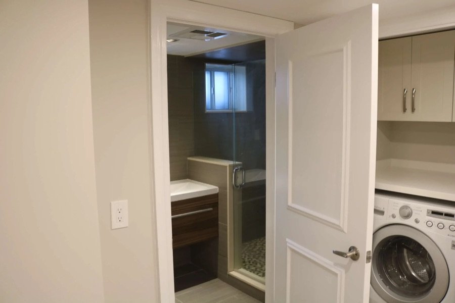 Basement renovation featuring a compact bathroom with glass shower enclosure and adjacent laundry area with front-load washer and storage cabinets