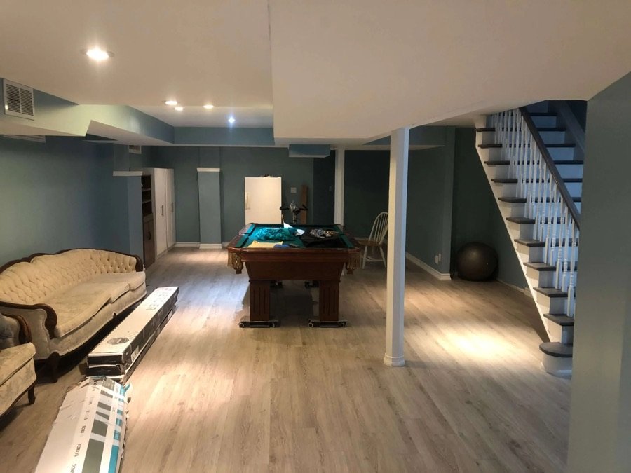 Renovated basement with pool table, light vinyl flooring, vintage sofa, and white staircase with open under-stairs space