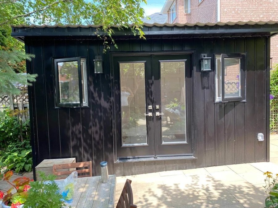 Fully finished backyard home addition featuring black vertical siding, double French doors, and new windows surrounded by patio and garden space