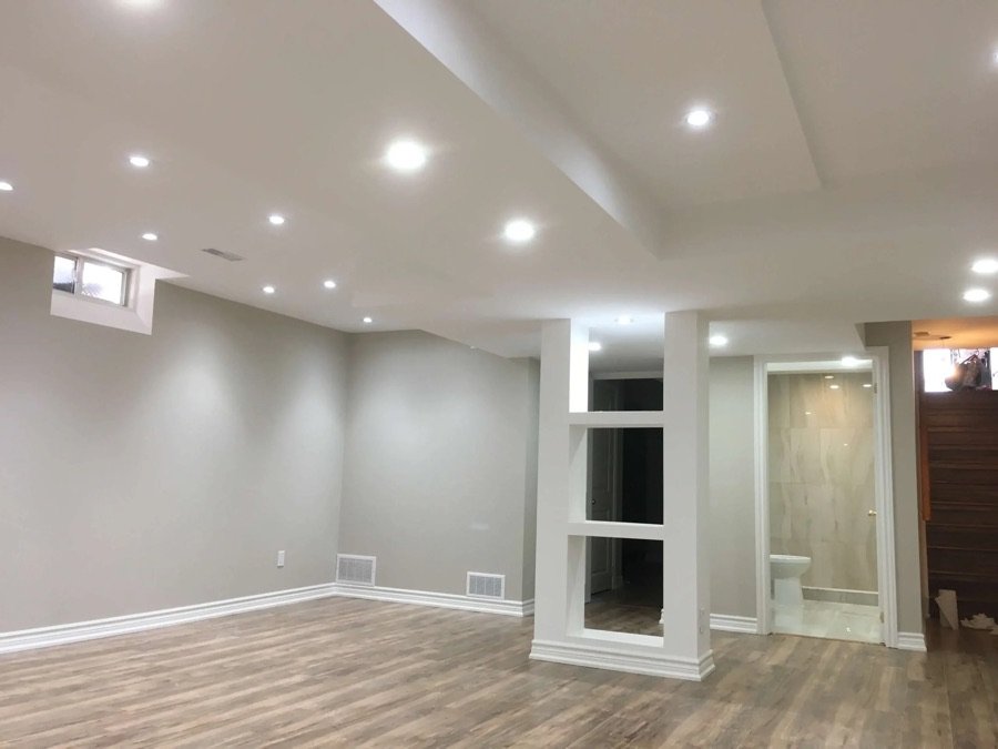 Spacious renovated basement with recessed lighting, neutral walls, custom shelving divider, and wood-look vinyl flooring adjacent to a modern bathroom