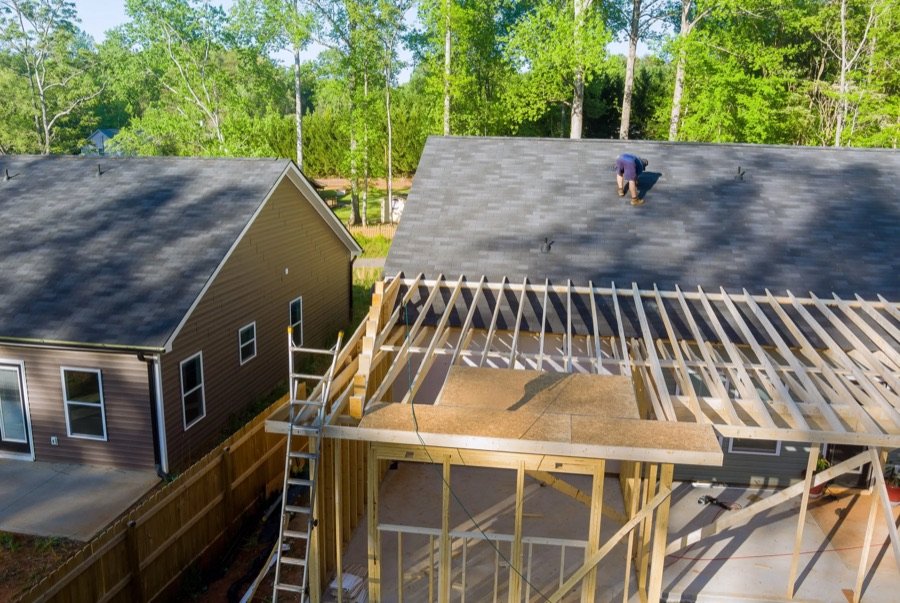 Residential home addition in progress with roof framing and plywood sheathing installation, showing attached extension under construction next to existing house