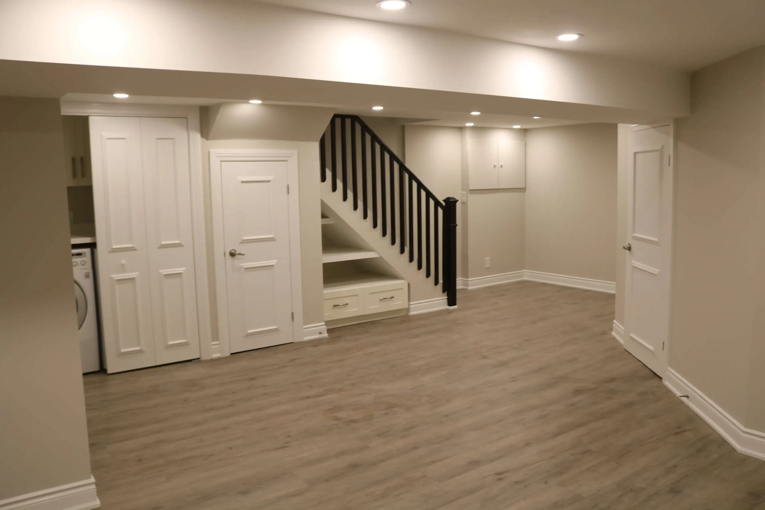 Renovated basement featuring light wood flooring, laundry closet, and built-in drawers with open shelving under the stairs