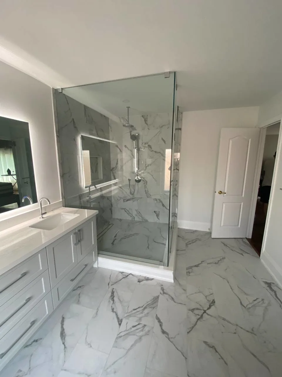 Bathroom renovation by RM Renovation showcasing marble-style tiles, frameless glass shower enclosure, and floating double vanity with LED mirror